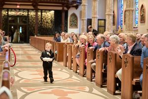 Wedding guests stand and watch as a couple walks down the aisle of an ornate church during a ceremony.