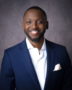 Professional headshot portrait of someone wearing a navy suit and white dress shirt against a gray studio background.