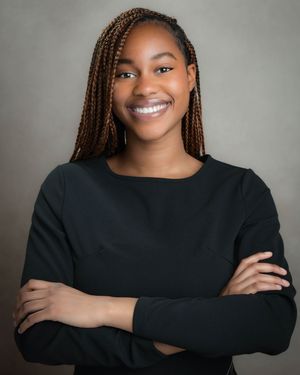 Professional headshot of a person with long braids wearing a black sweater, smiling warmly with arms crossed.