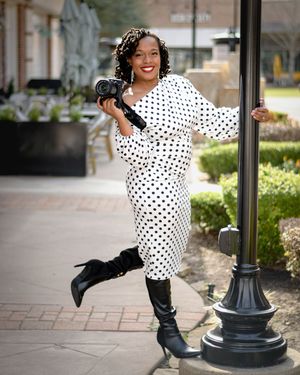 Woman in polka dot dress and black boots poses playfully with a light post in an outdoor setting.