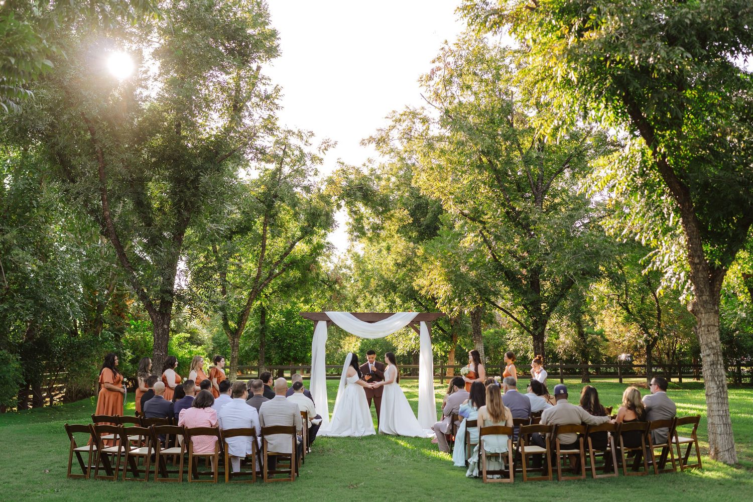 Two brides stand beneath an arbor during their wedding ceremony, surrounded by family and friends in a green grove of pecan trees