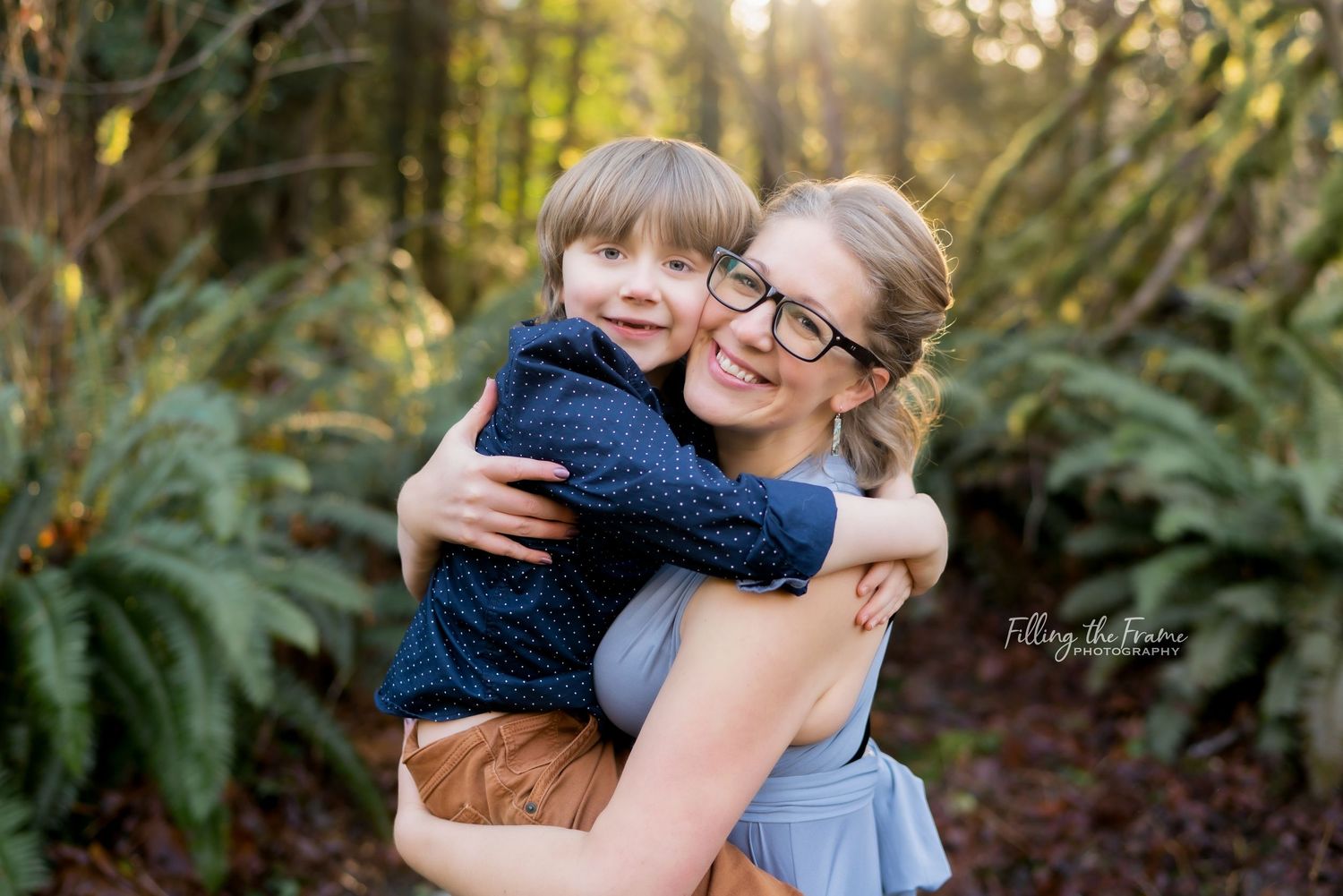 A mother and child share a joyful embrace outdoors in a wooded setting during golden hour.