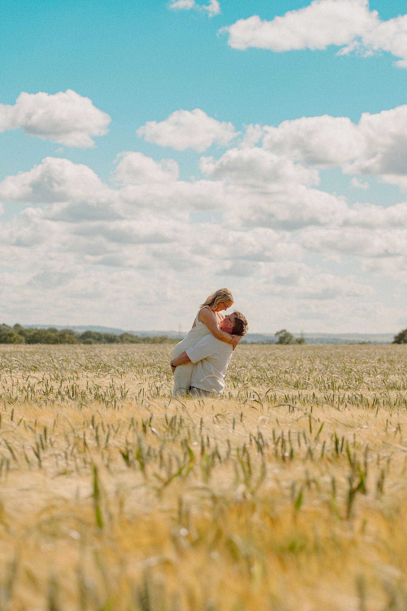 Couple embracing in a golden wheat field under a bright blue sky with fluffy white clouds on a sunny day.