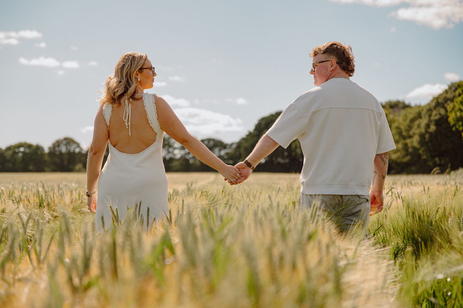 Couple holding hands in a golden wheat field on a sunny day, woman in backless white dress.