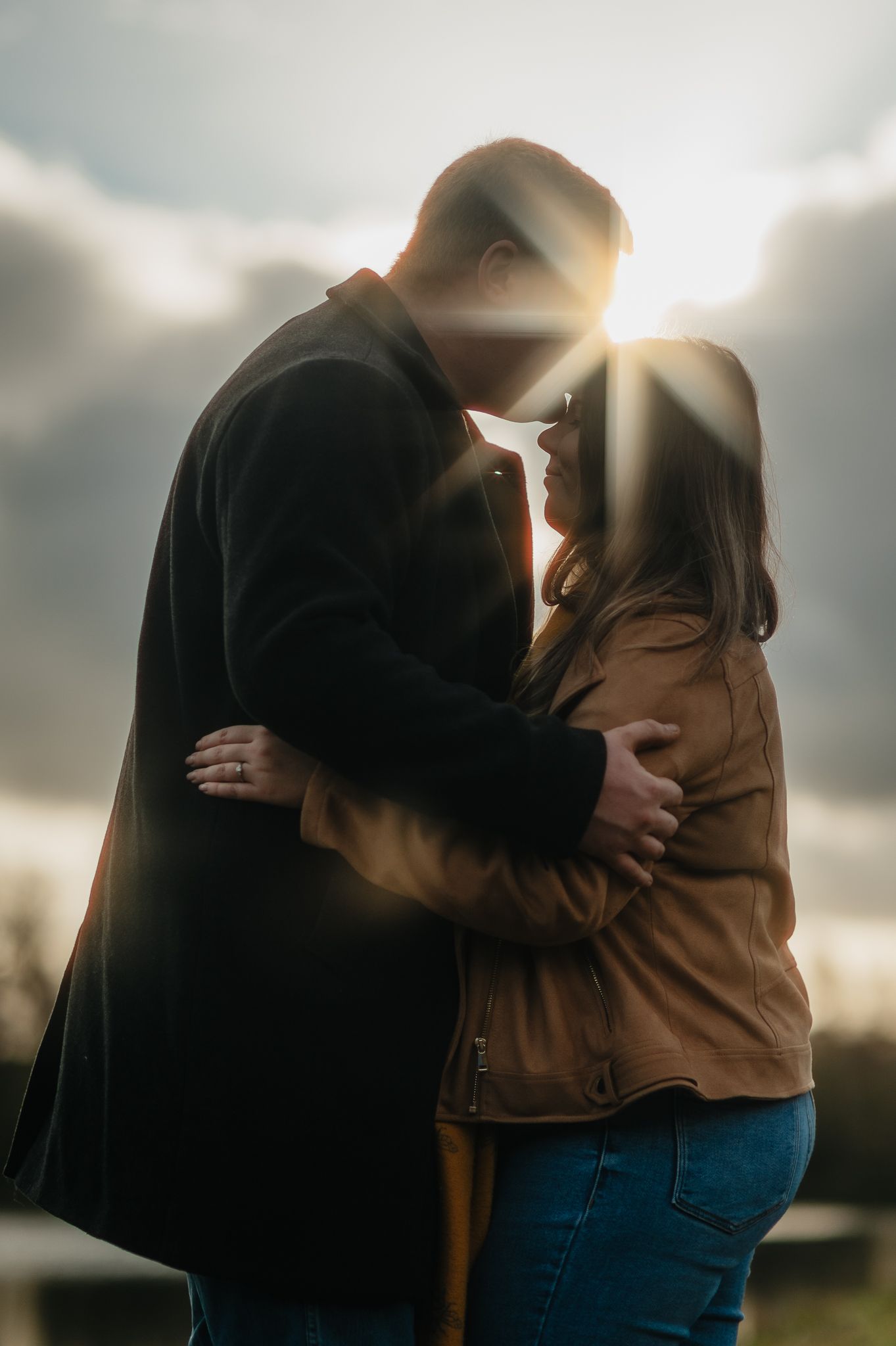 A couple embraces in silhouette against a cloudy sky with sunlight peeking through during sunset.