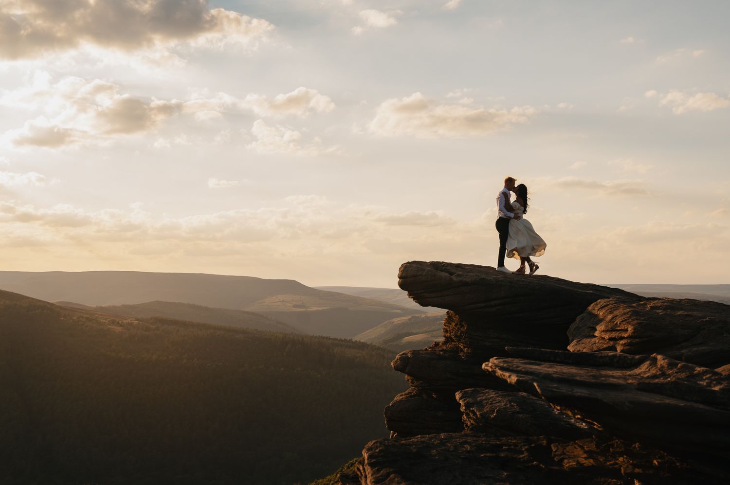 Two silhouettes stand on a dramatic cliff edge overlooking mountains during a golden sunset.