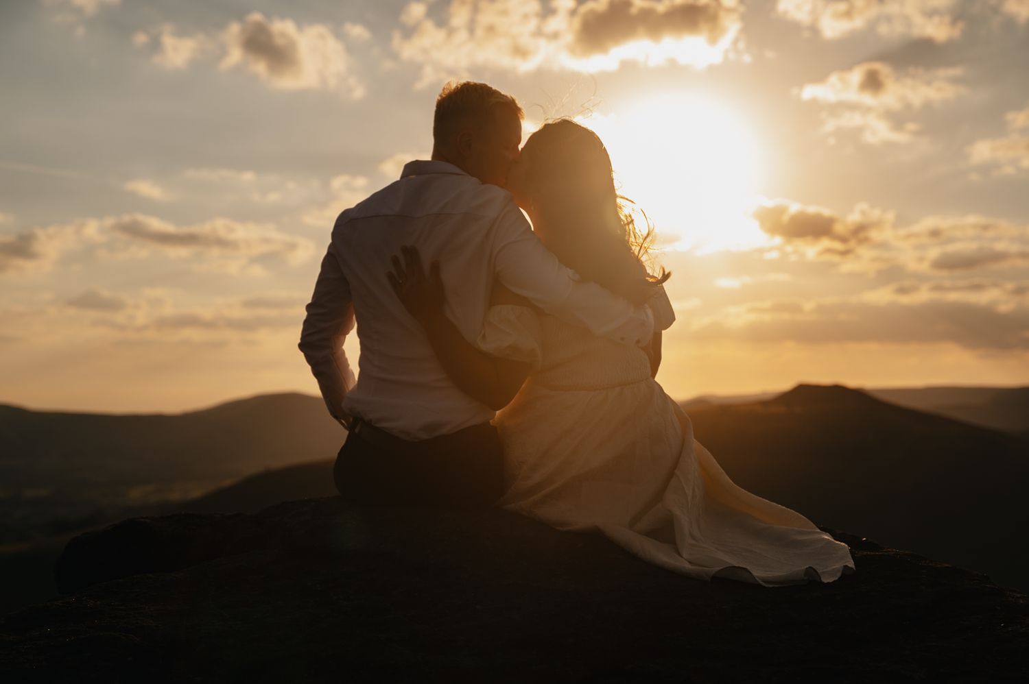 A silhouetted couple embraces against a dramatic sunset sky with mountains in the background.