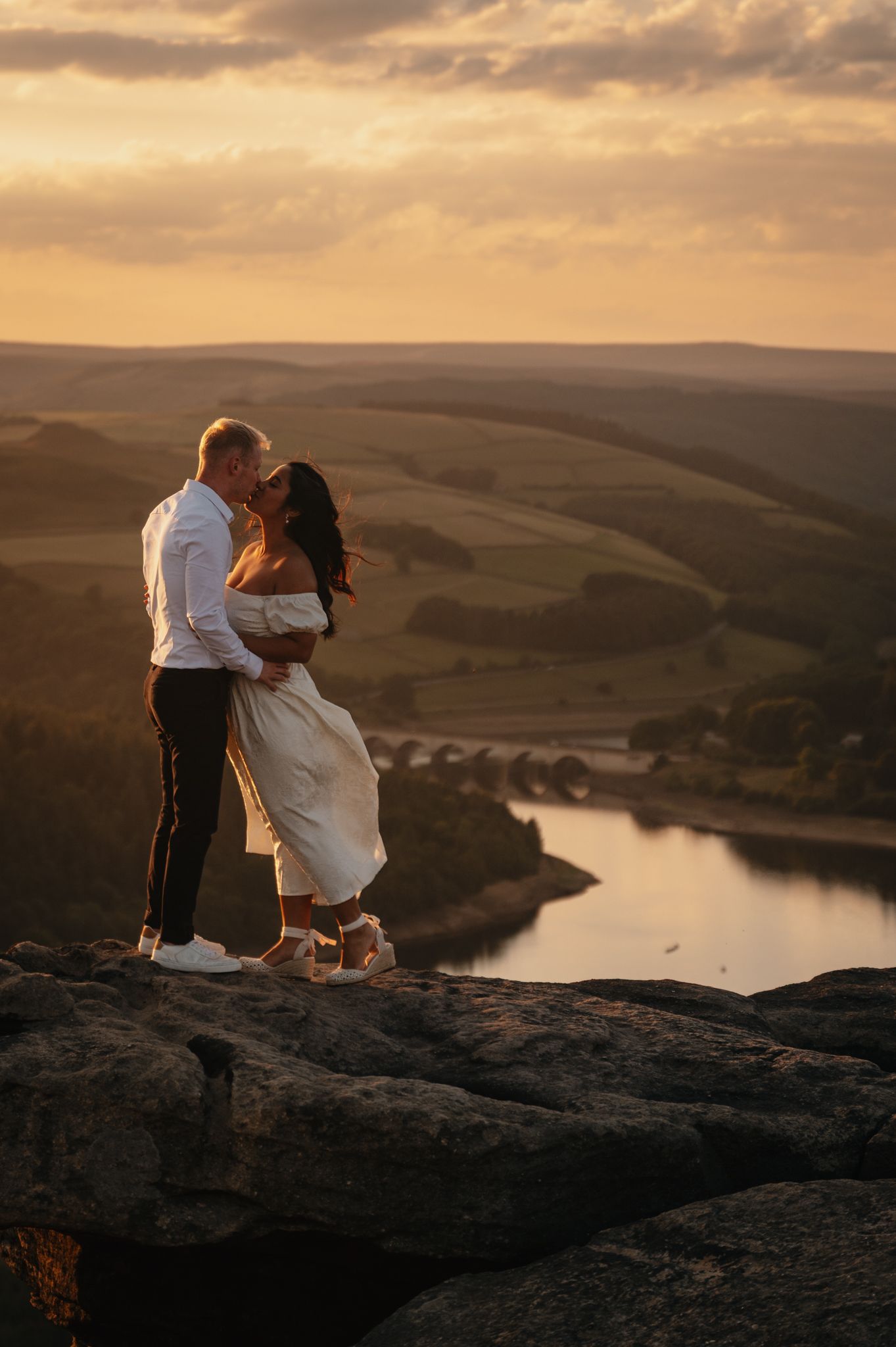 Two people embrace on a rocky cliff overlooking a scenic reservoir at sunset with dramatic clouds in the sky.