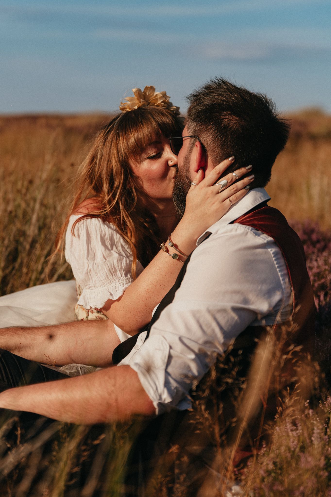 Couple sharing a romantic kiss in a golden field, woman wearing white dress and floral hair accessory.