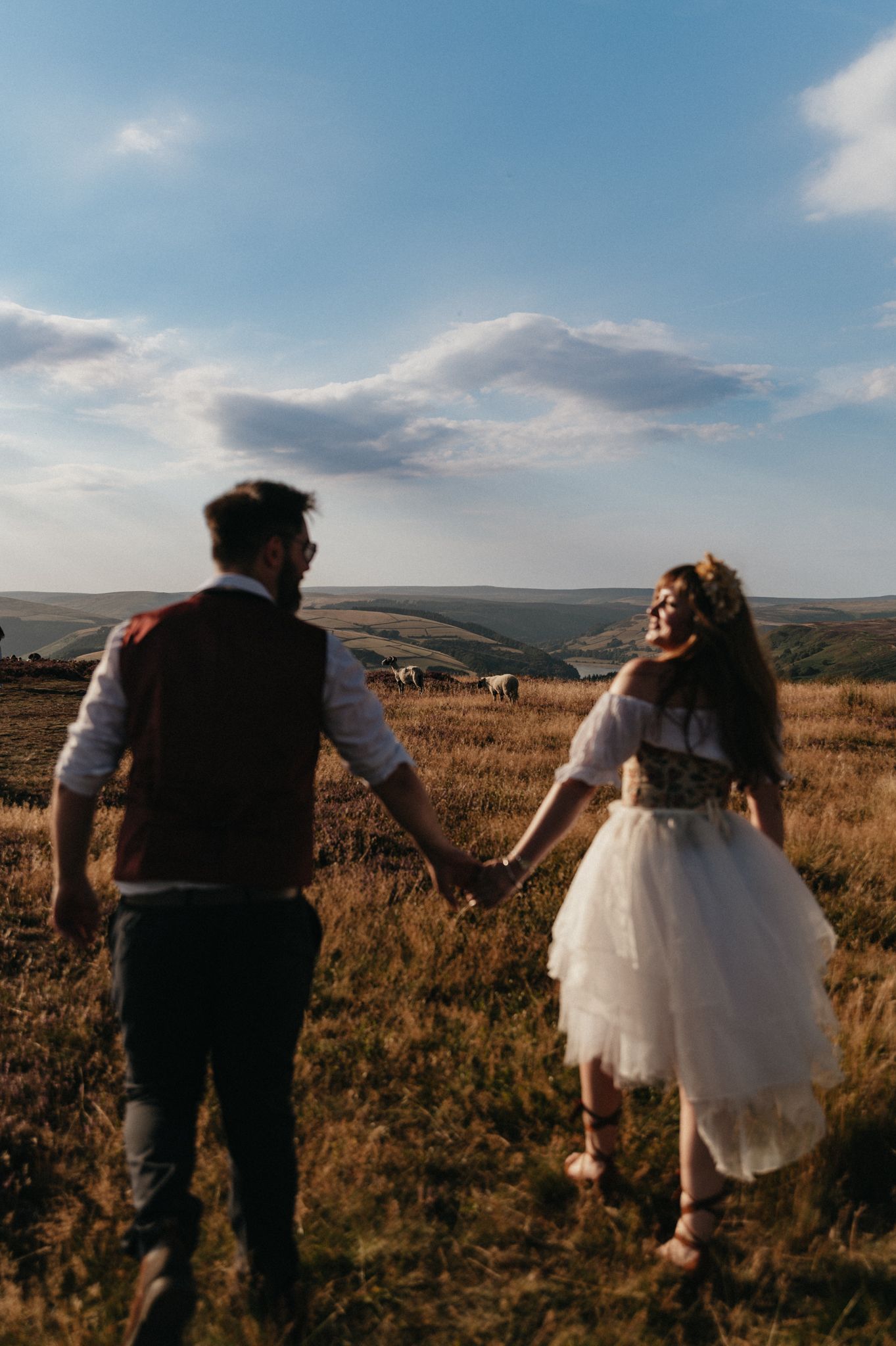 A bride and groom hold hands walking through golden fields under a blue sky on their wedding day.
