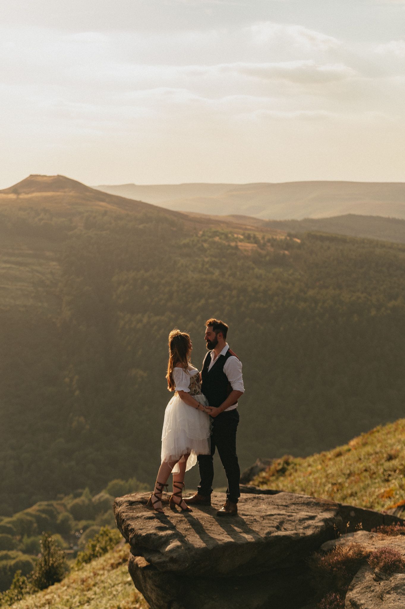 Couple in wedding attire standing on a rocky cliff at golden hour with scenic mountain forest views.