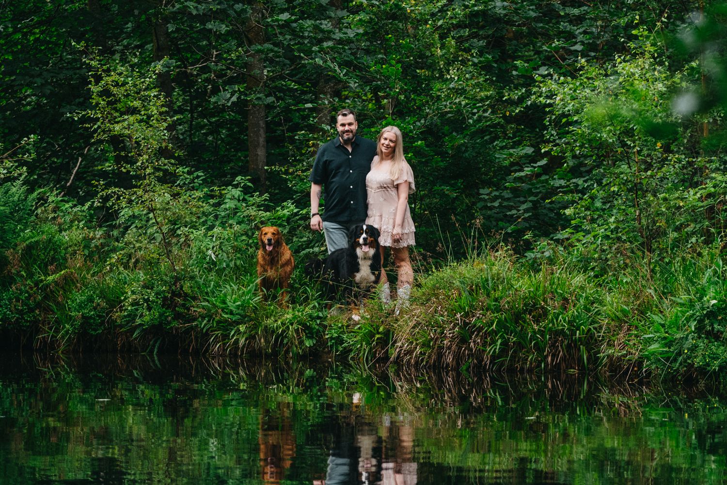 A couple and their dog stand along a serene forest pond with lush greenery reflecting in the calm water.