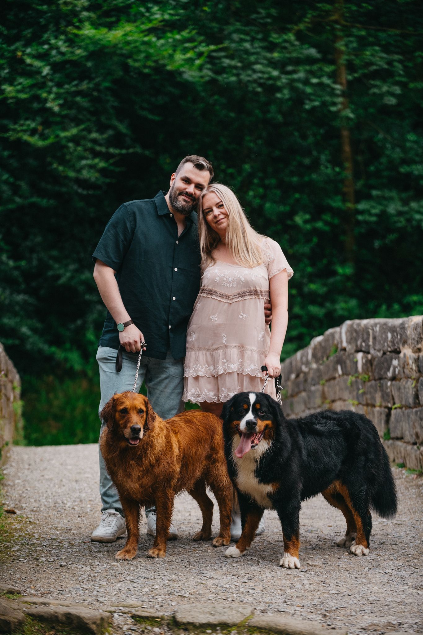 A couple poses with their two dogs on a stone path surrounded by lush green foliage in the background.