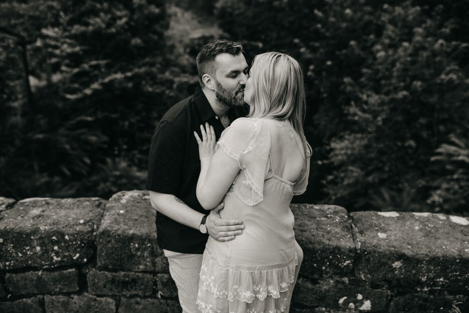 A romantic black and white engagement photo of a couple embracing on a stone wall in a forest setting.