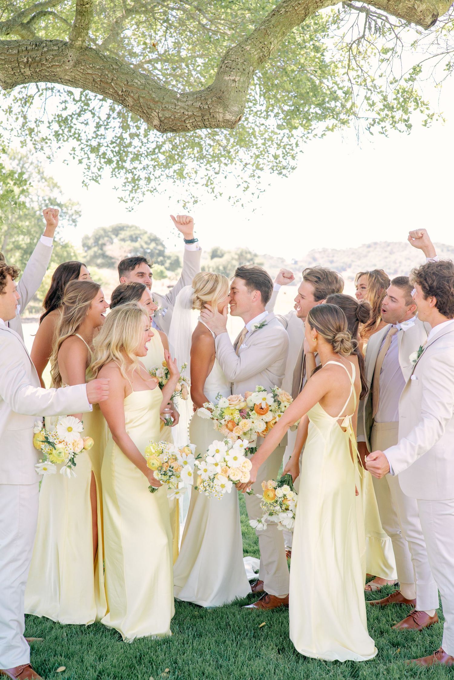 Wedding party in yellow dresses and white attire celebrates under trees during outdoor ceremony.