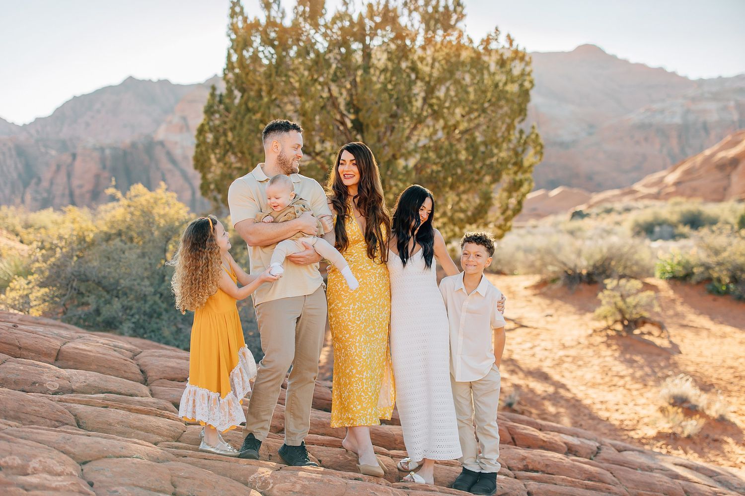 Family group wearing coordinated yellow and white outfits poses together in a scenic desert mountain landscape at sunset.