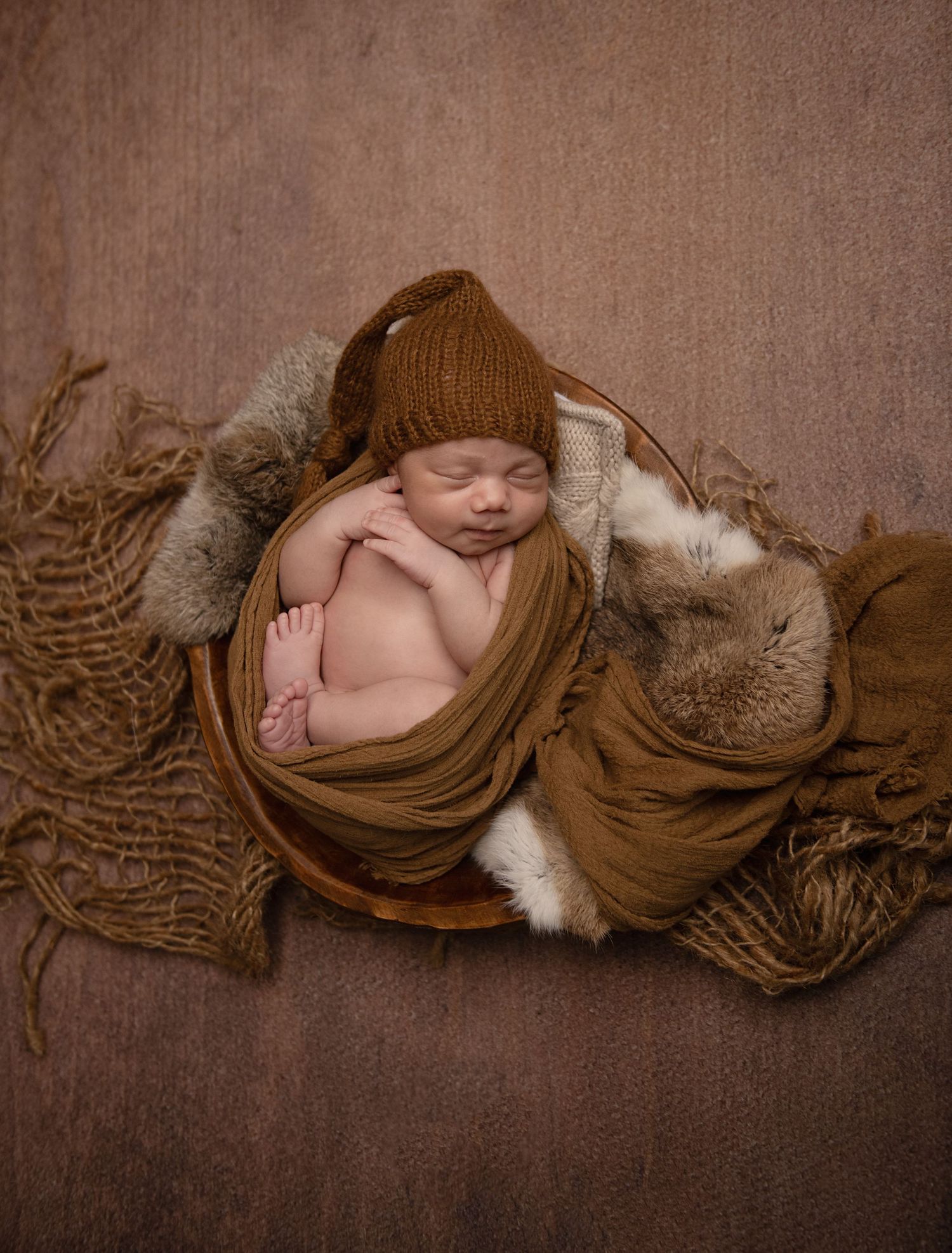 Sleeping newborn wrapped in brown fabric and knit hat nestled in cozy basket with textured fabrics and fur accents.