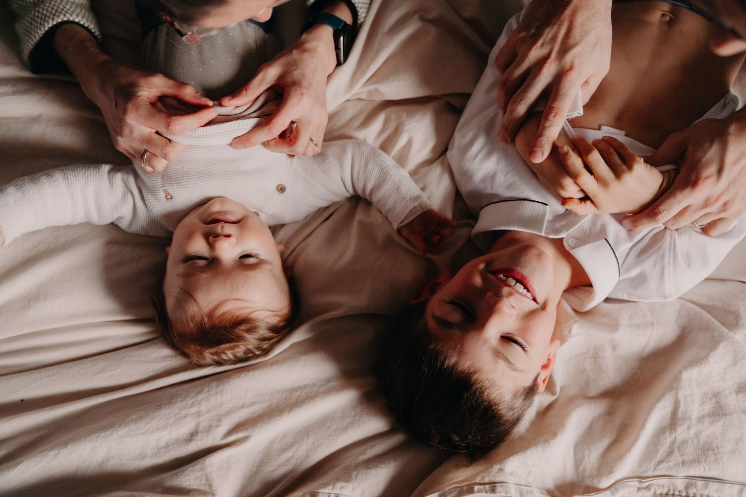 Two babies lay side by side on white bedding while parents playfully interact.