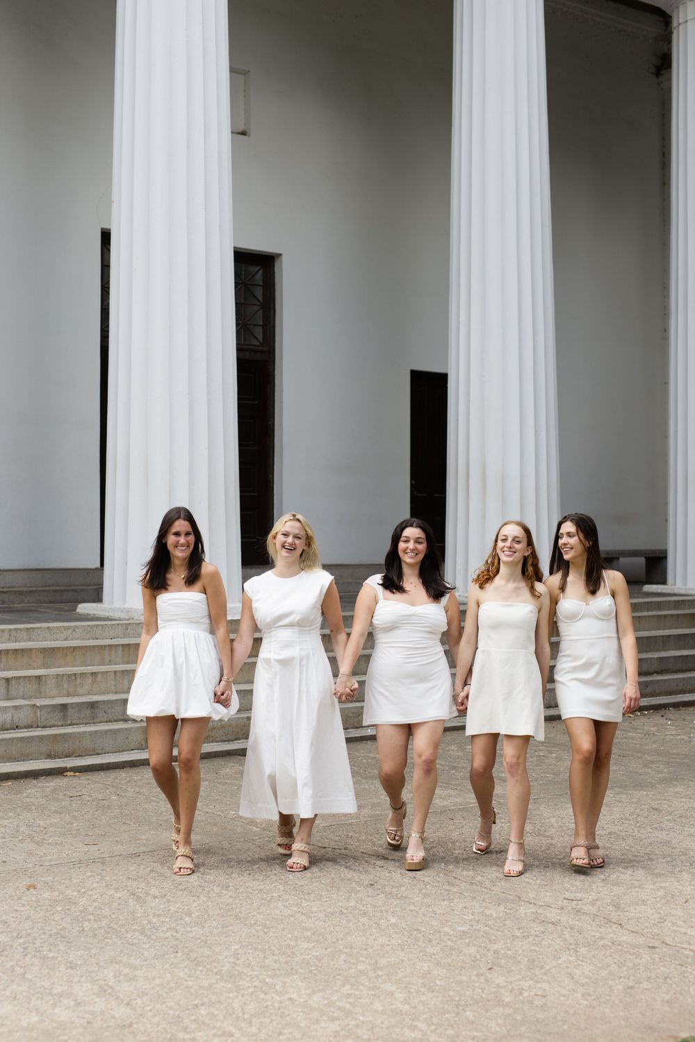 Group of graduates in white dresses pose together in front of classical columns.