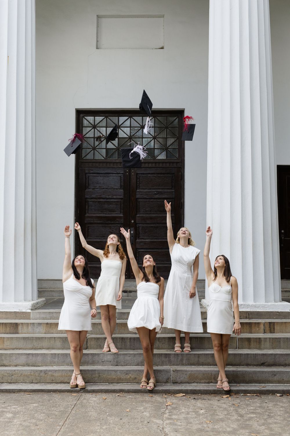 Graduates in white dresses celebrate by throwing caps in front of columned building.