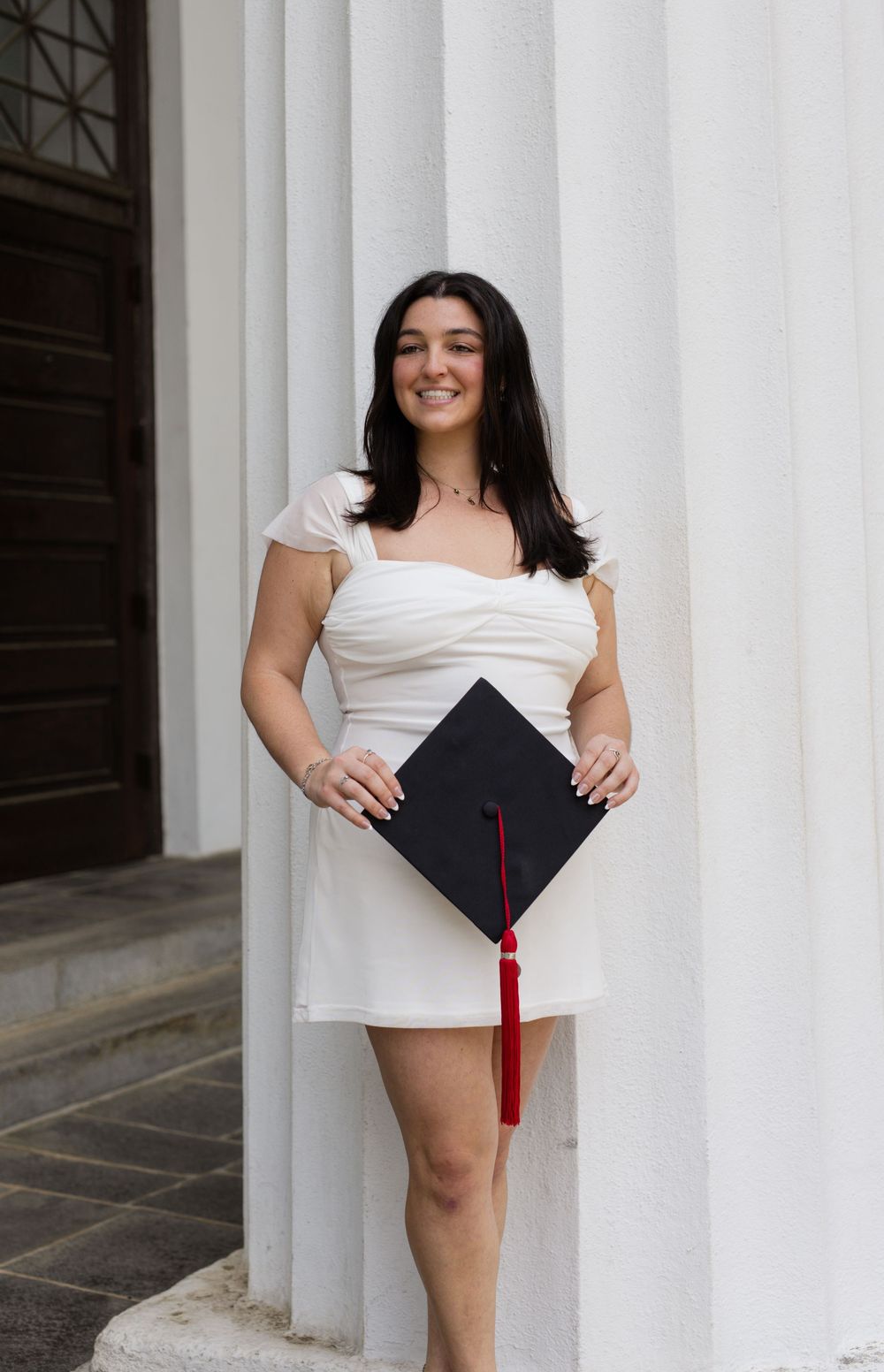 Graduate in white dress poses playfully with cap against architectural columns.