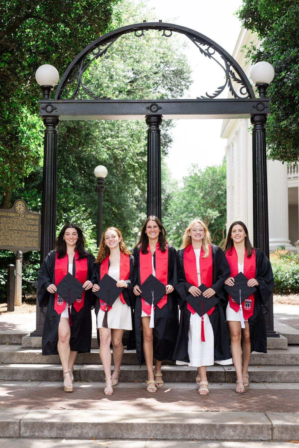 Graduates in red and black attire pose under decorative arch on campus steps.