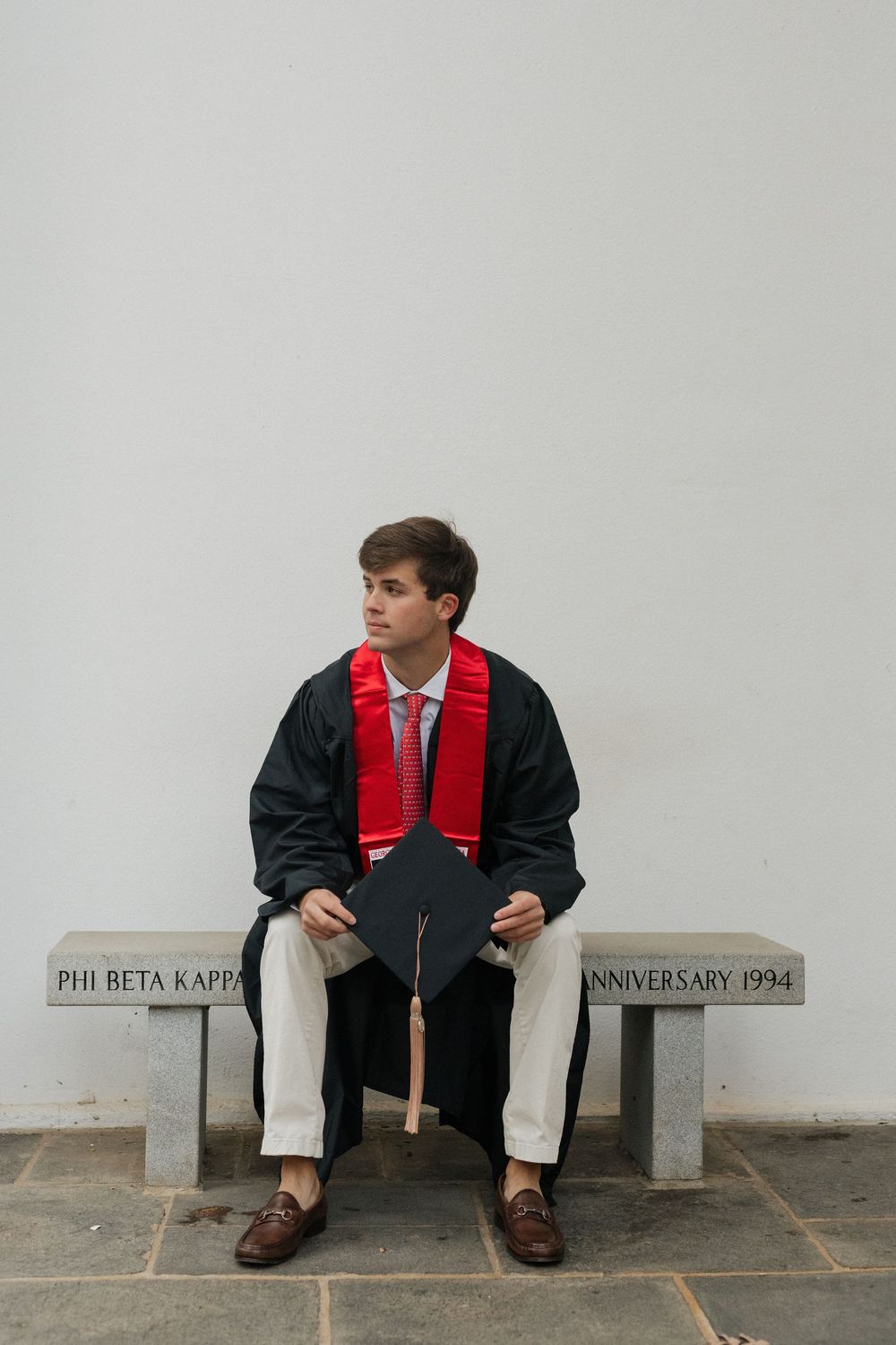 Graduate in black gown and red stole sits on Phi Beta Kappa bench in formal attire.