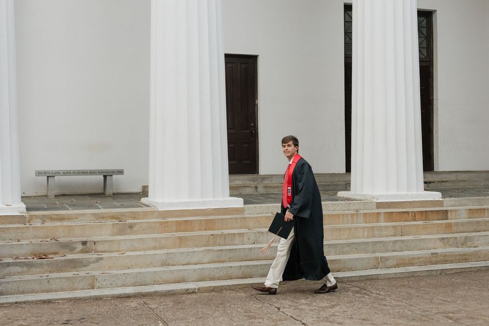 Graduate in academic regalia walks past white columns of classical building.