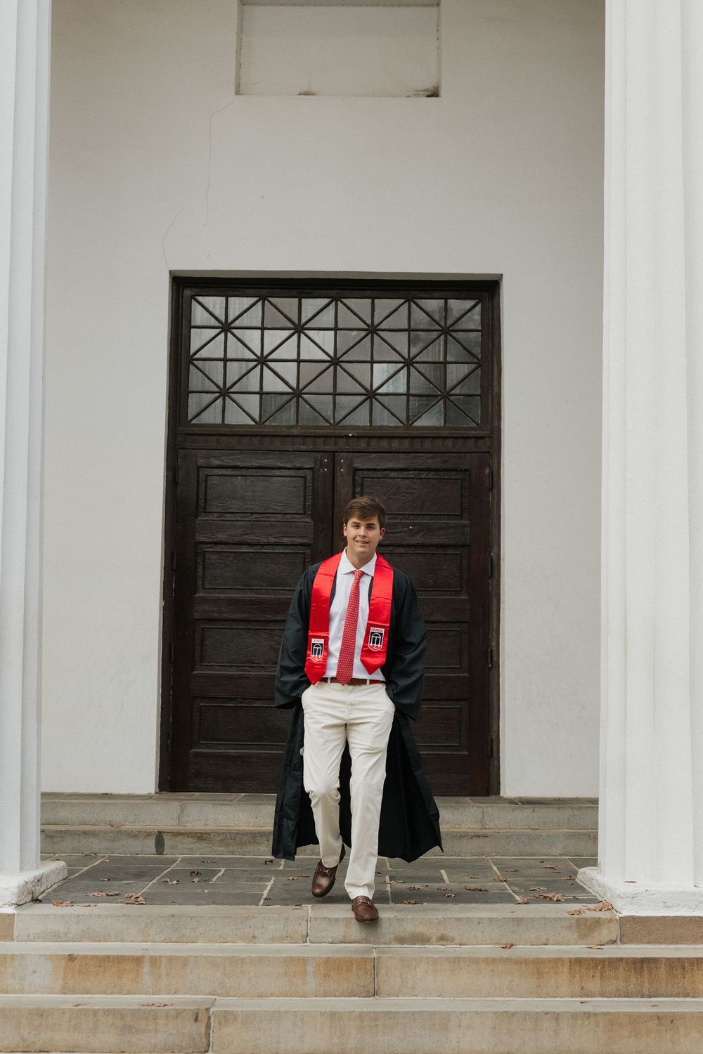 Graduate in cap and gown poses in front of ornate dark wooden door with geometric patterns.