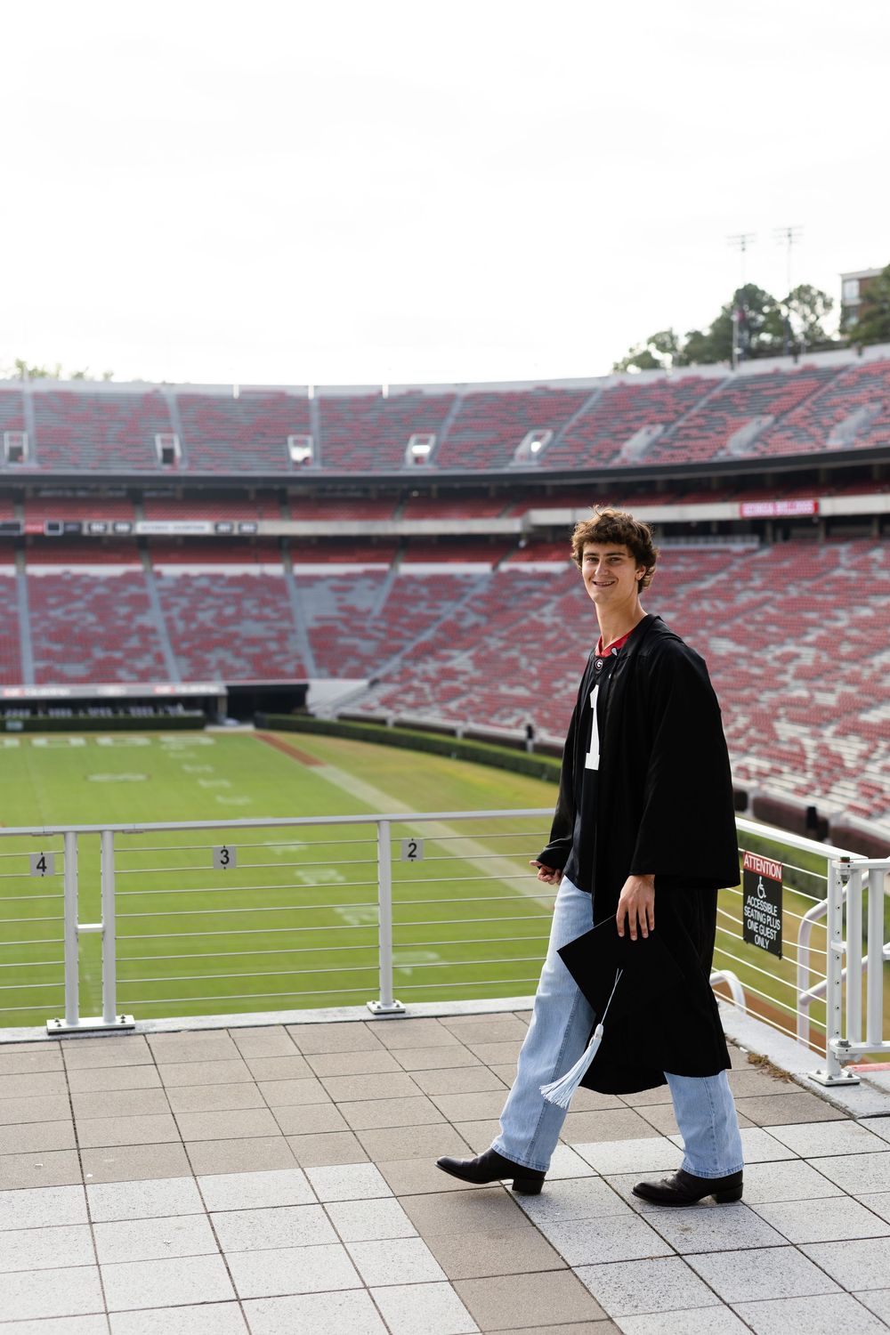 Graduate in black academic gown stands on stadium viewing deck overlooking football field.