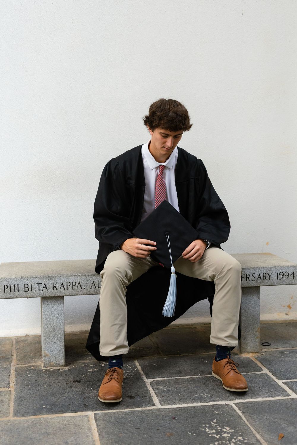 Graduate in black gown holds blue tassel while sitting on stone bench with Phi Beta Kappa inscription.