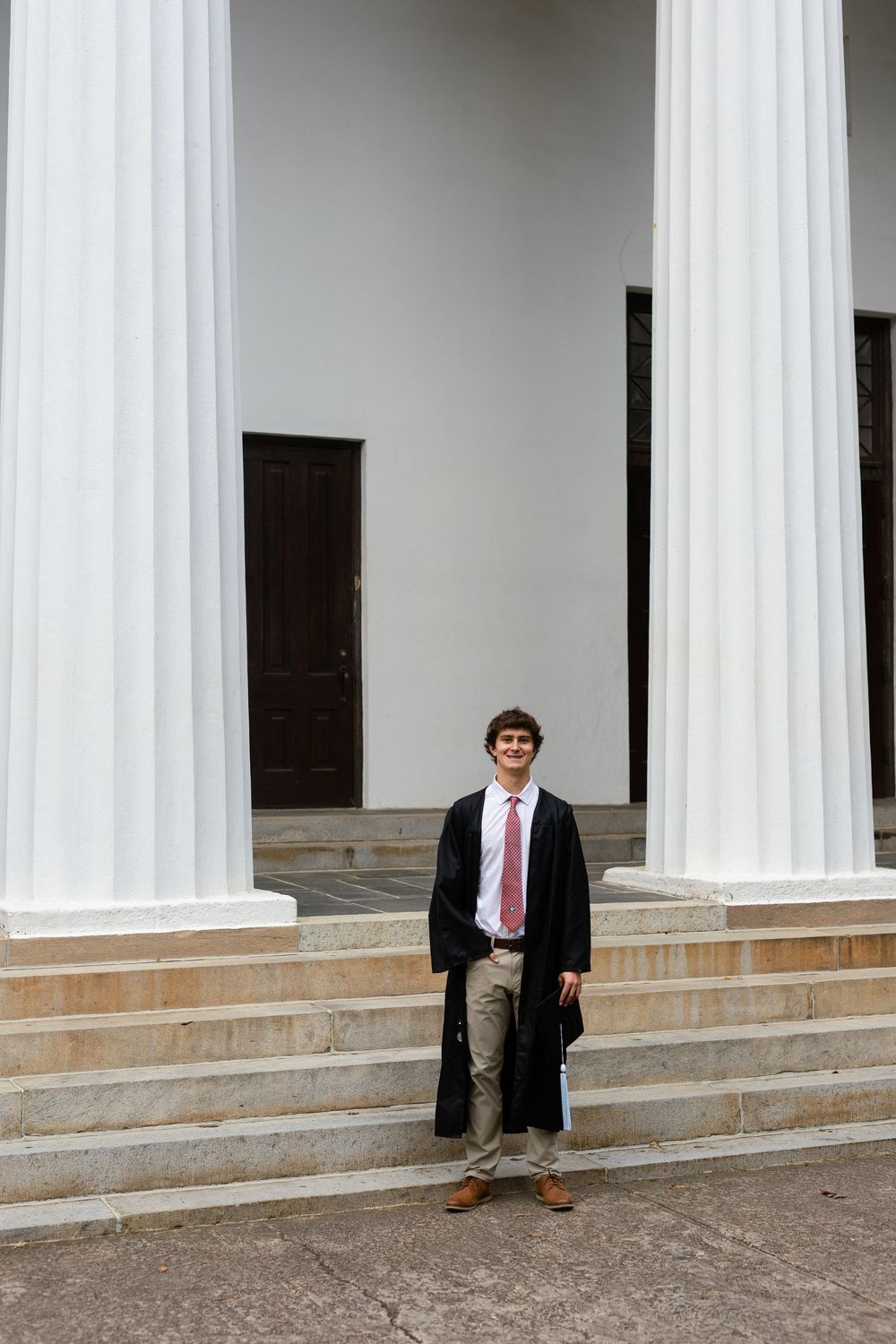 Graduate poses on steps between white columns in academic regalia and khaki pants.