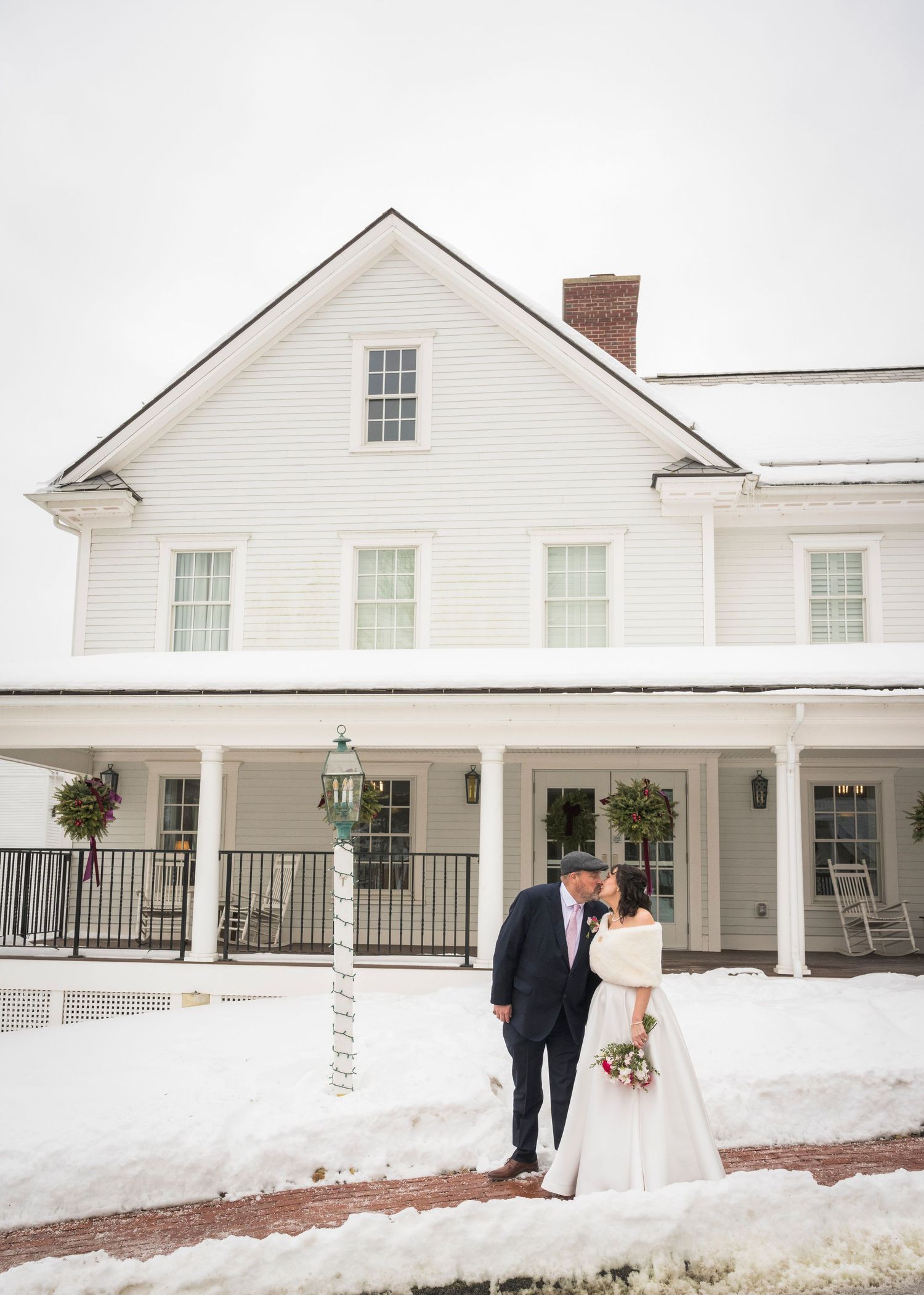 Winter wedding panorama at a white colonial-style venue with snow-covered grounds and long covered porch.