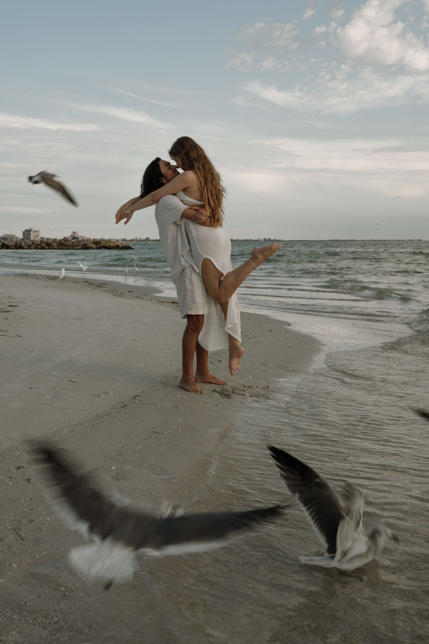 A romantic beach scene with two figures embracing on the shore while seagulls fly past at sunset.