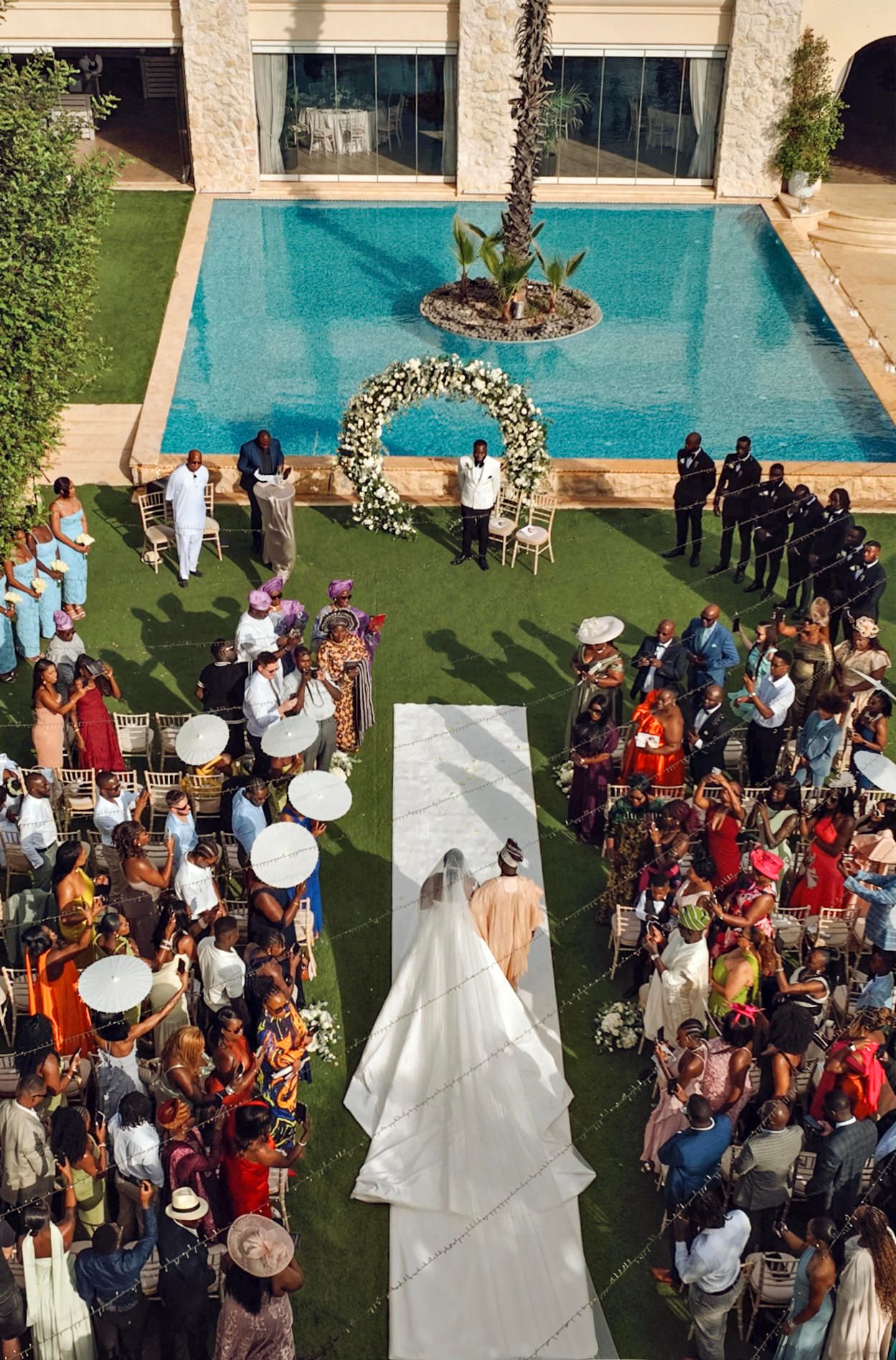 Outdoor wedding ceremony by a pool with guests seated on chairs and palm trees in the background.