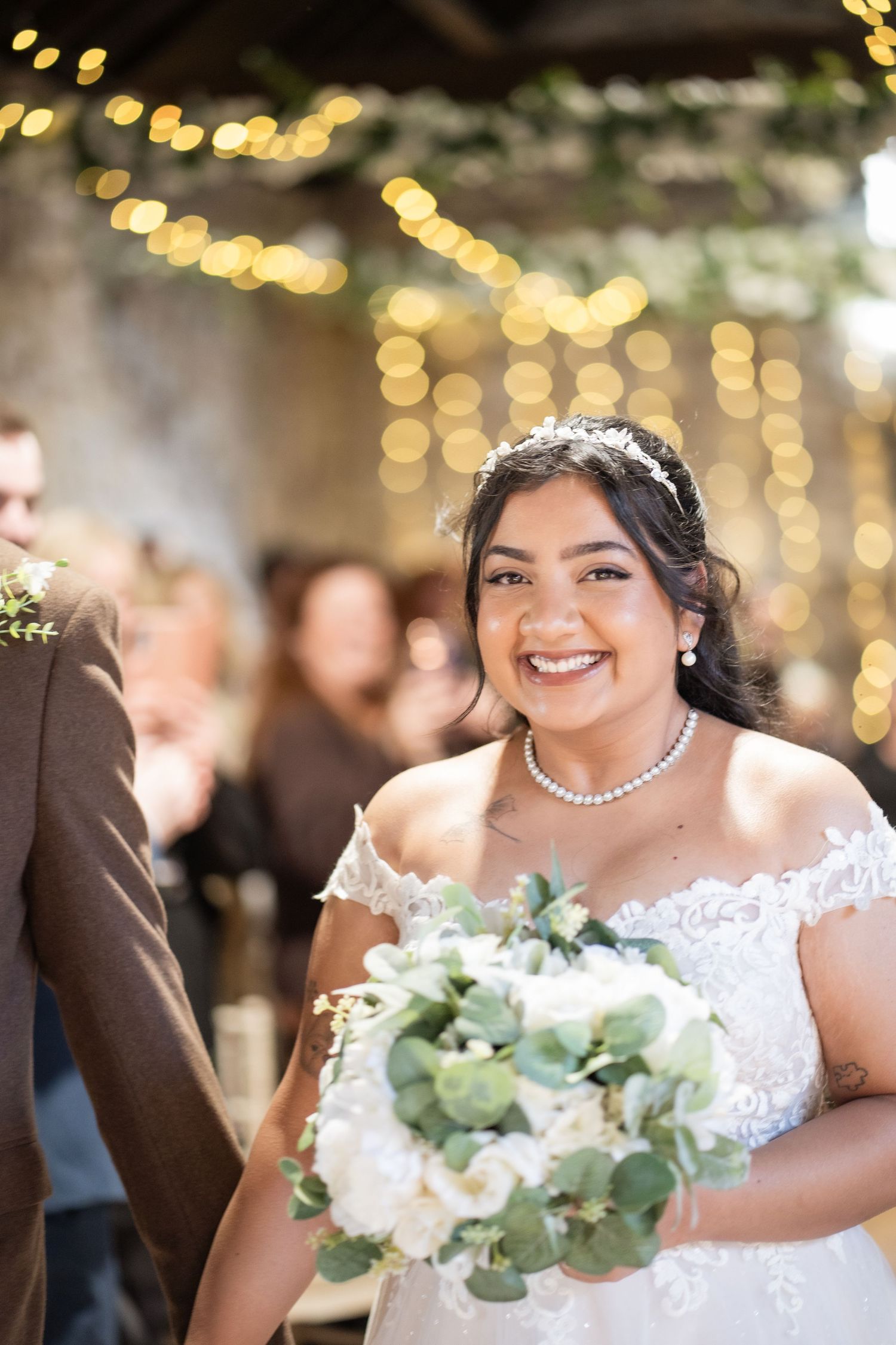 Beautiful bride in off-shoulder lace dress holds white and green bouquet with twinkling lights in background.