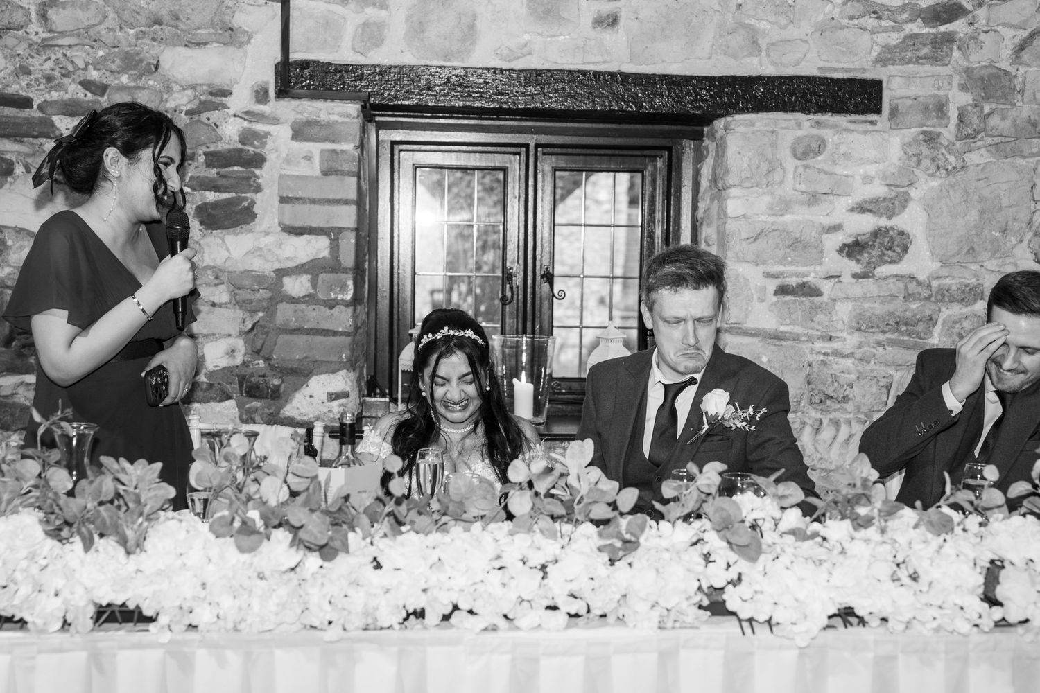 Wedding guests seated at long tables with stone walls and floral centerpieces during an indoor reception celebration.