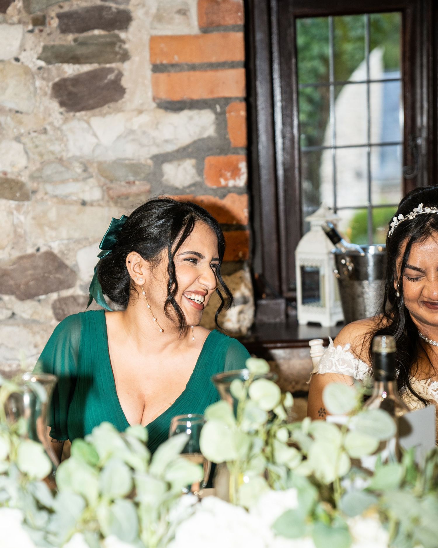 Wedding party members in emerald green dresses share joyful laughter together by a rustic stone wall.