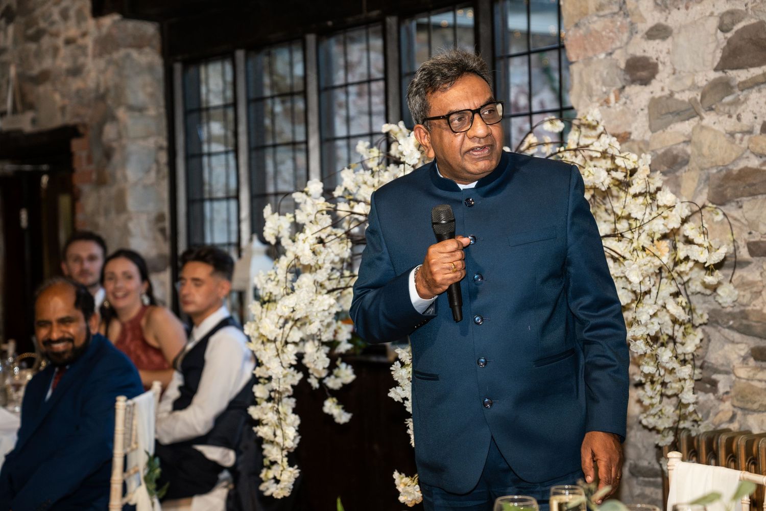 A speaker addresses guests at an elegant wedding reception under a floral arch against a stone wall.