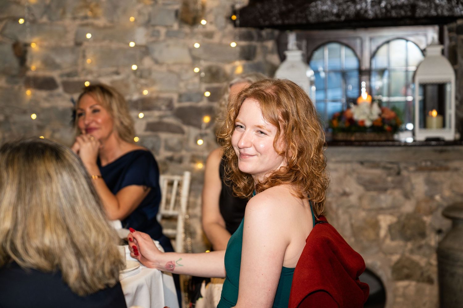 A person in a green dress smiles at a festive indoor gathering with string lights and stone walls.
