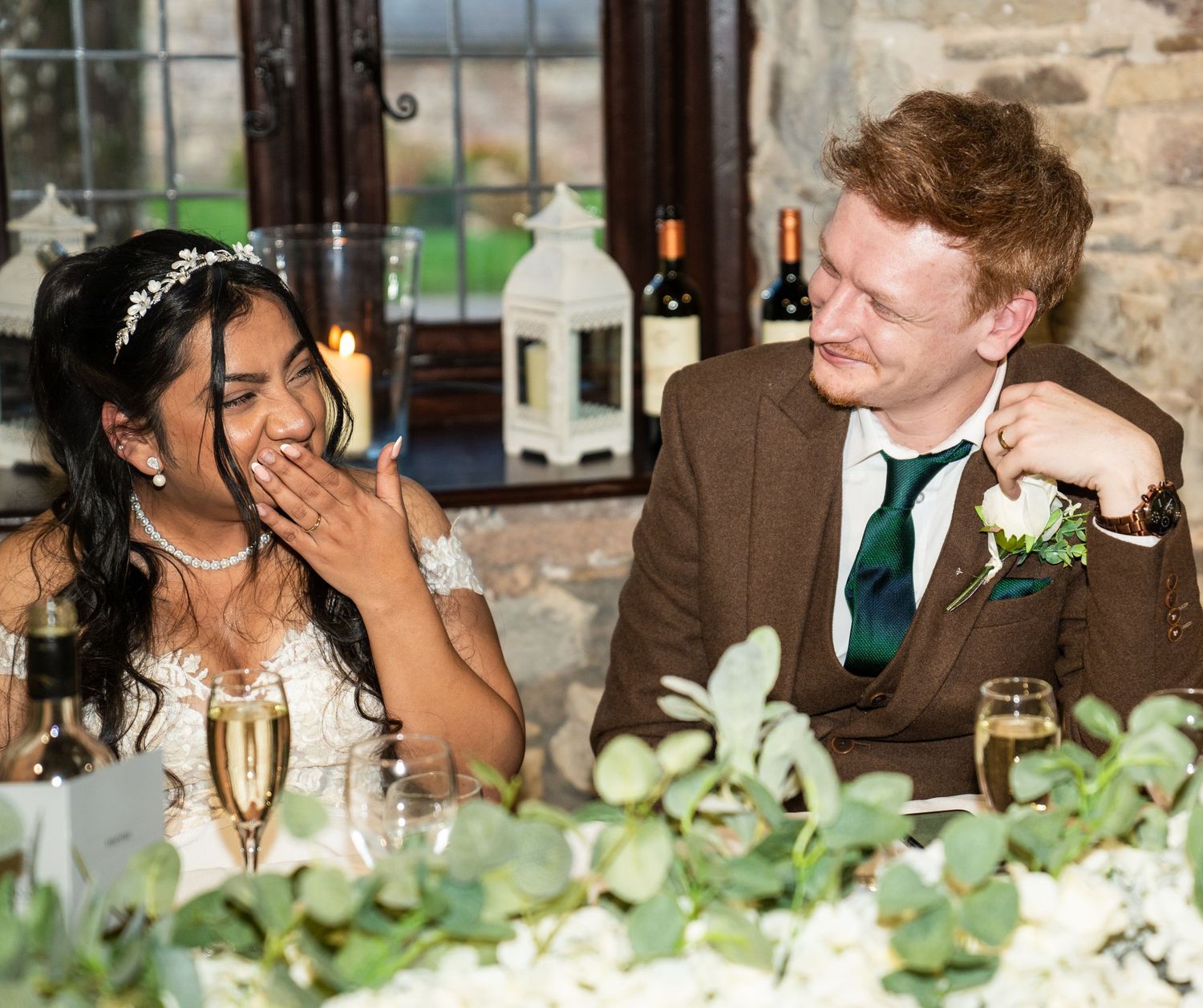 Wedding reception guests share joyful laughter while seated at a table decorated with greenery centerpieces.