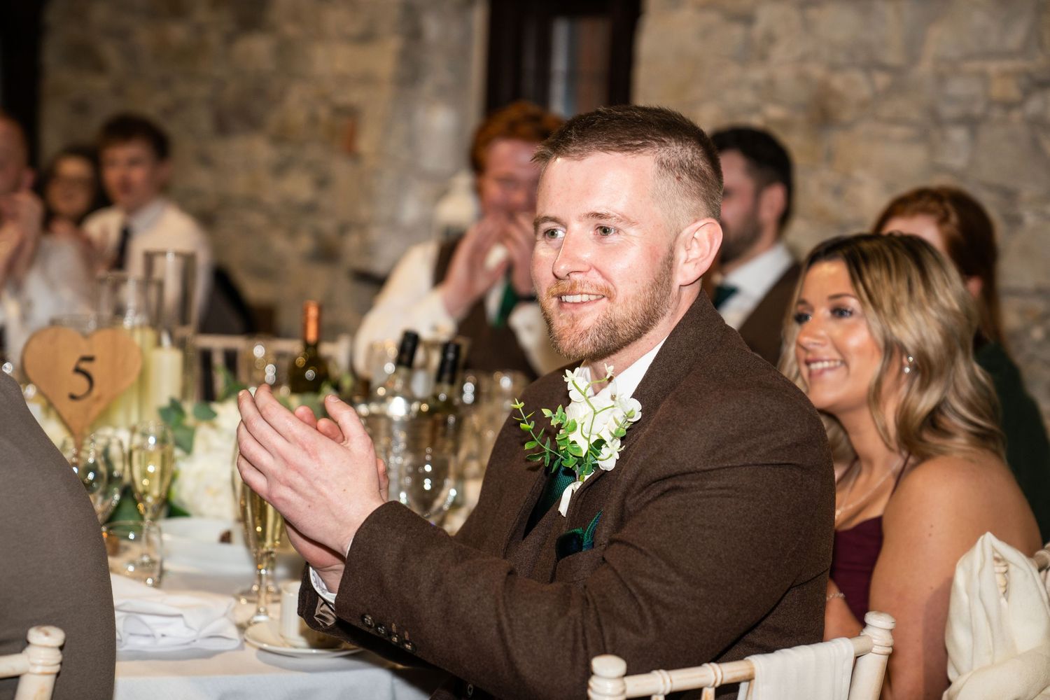 Wedding guests laugh and smile during a reception dinner in a rustic stone-walled venue.