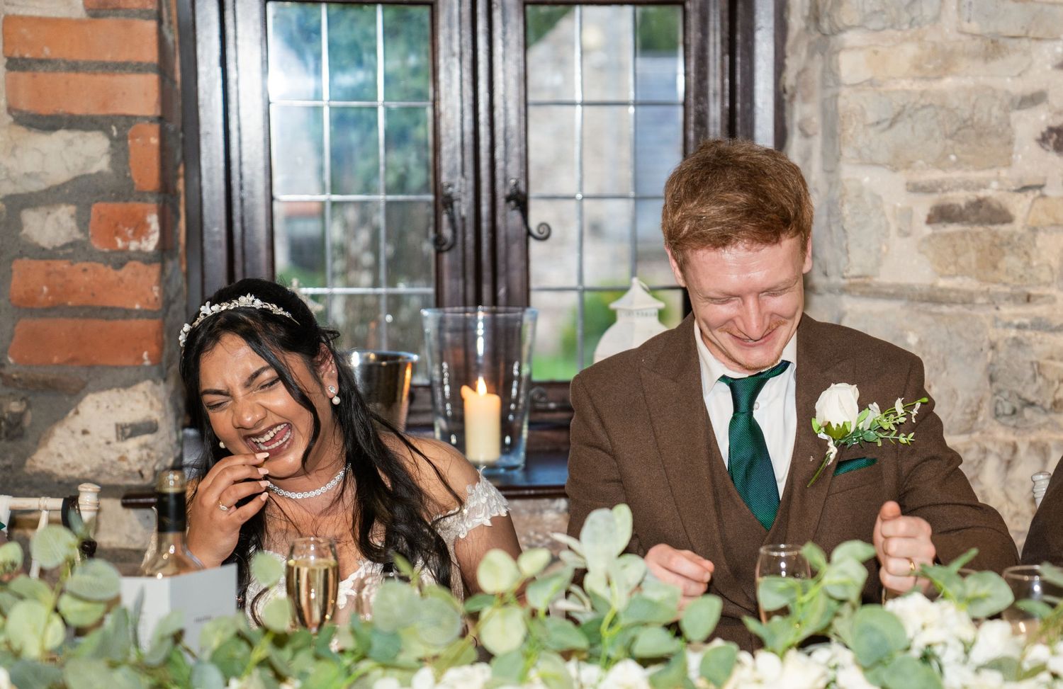 A couple shares a joyful moment while seated at their wedding reception table decorated with greenery.
