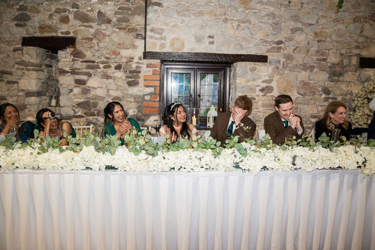 A panoramic view of guests seated at a long table during an outdoor dinner party against a stone wall backdrop.