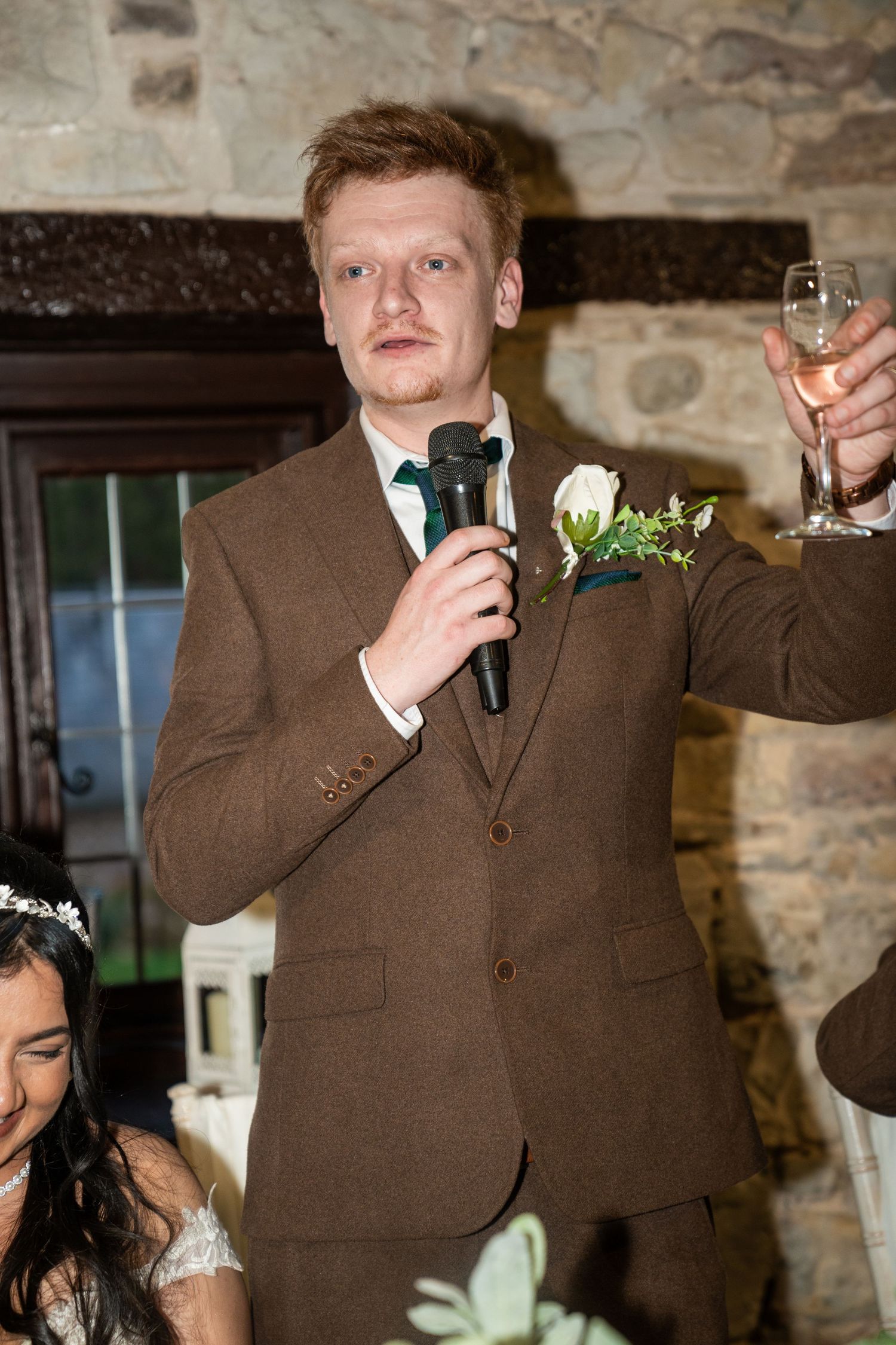 Wedding guest in brown suit delivers speech with microphone at rustic stone venue during reception celebration.