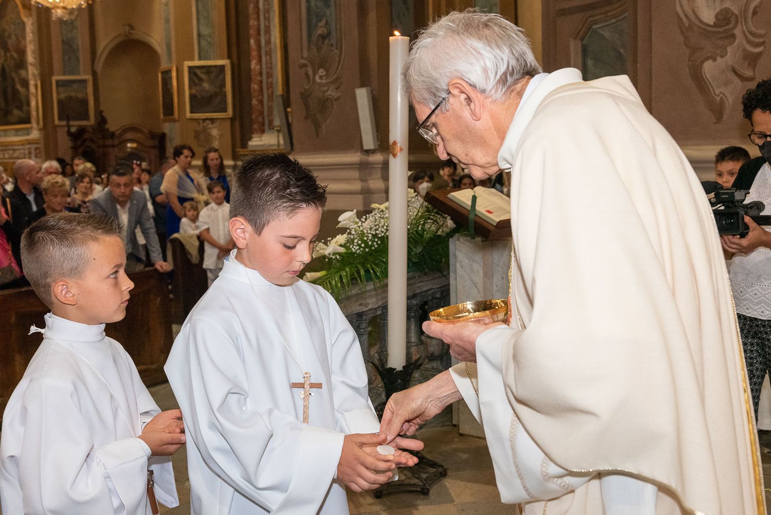 Priest in white vestments presenting items to young altar servers during church service.