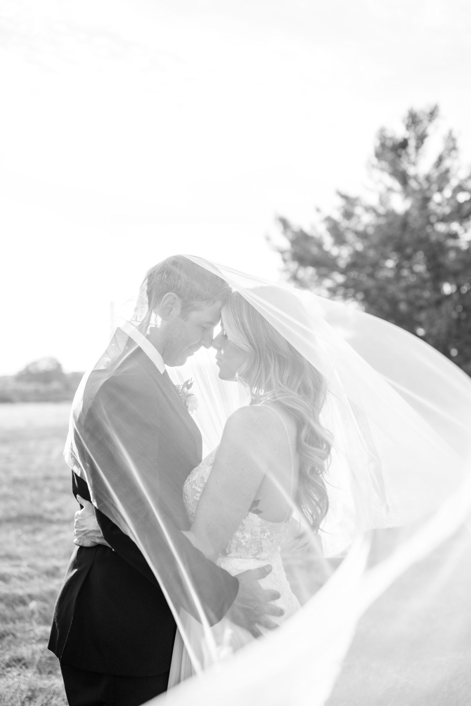 A romantic black and white wedding photograph showing a couple embracing as a delicate veil flows in the wind.