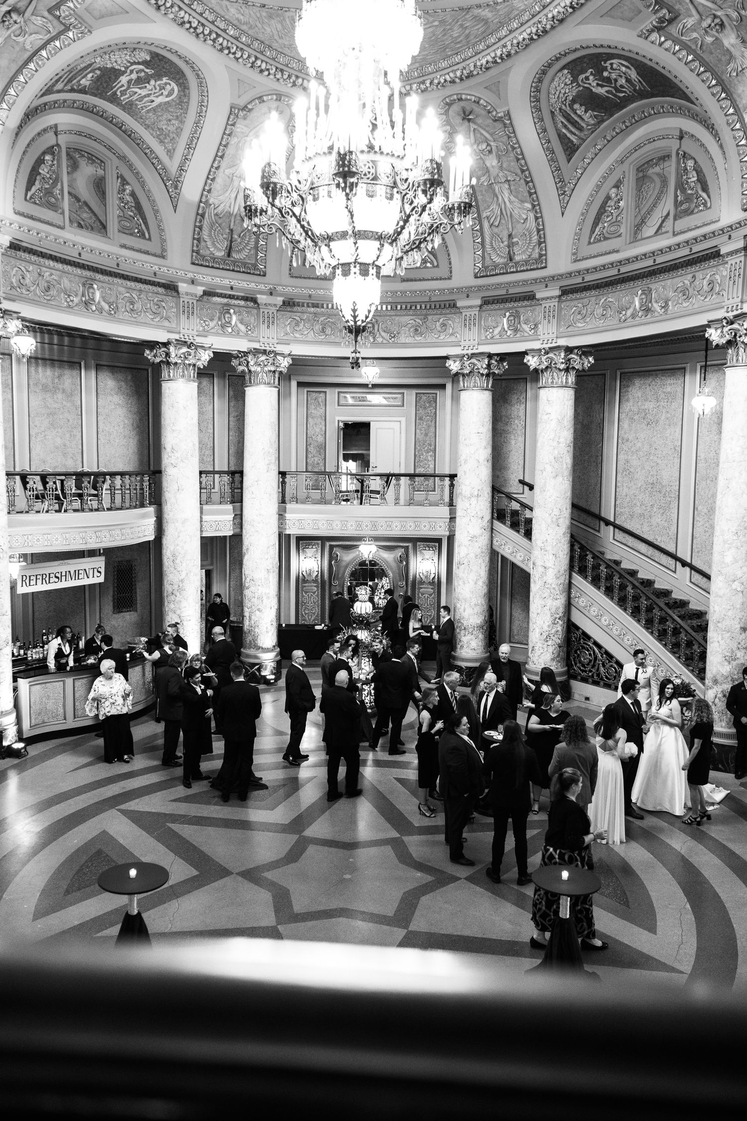 Elegant black and white panoramic view of guests mingling in an ornate ballroom with crystal chandeliers and arched ceilings.