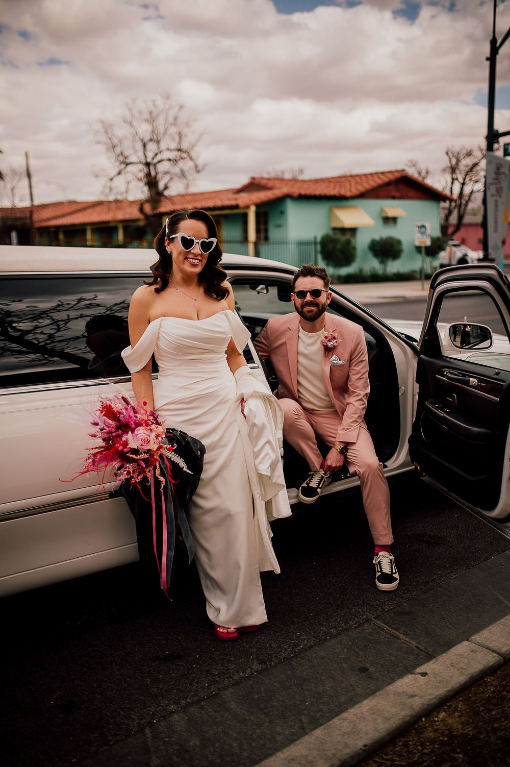 A couple exits their wedding car, with the bride in white holding a pink bouquet and the groom in a pink suit.