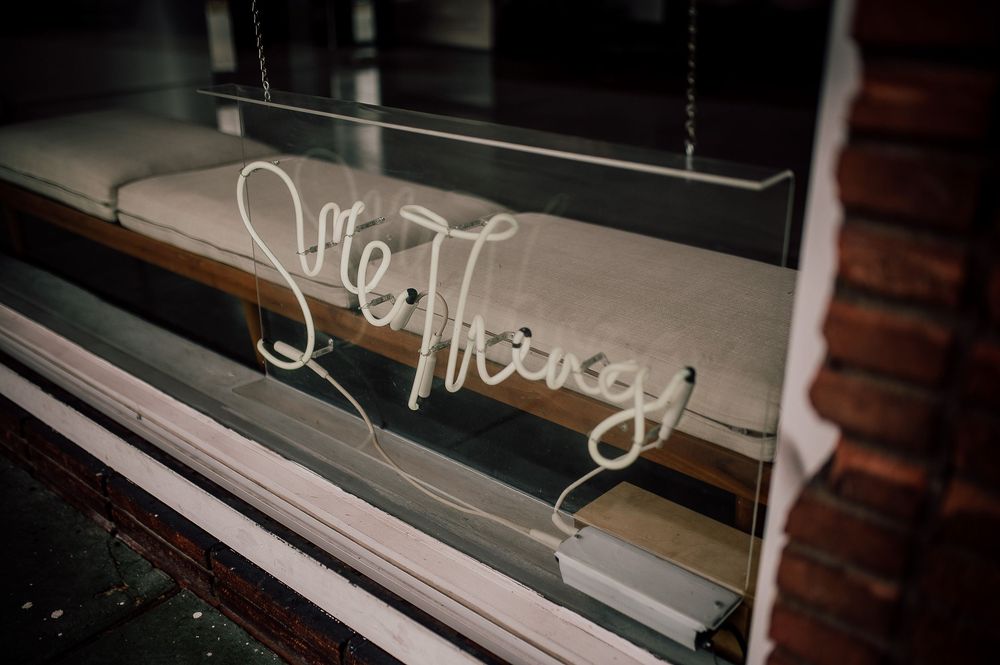 A neon sign reading 'She/They' glows softly in a window display at night.
